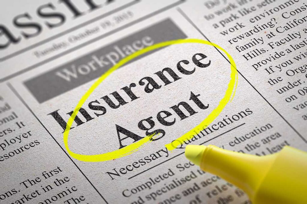 A middle-aged person sits at a kitchen table sorting through insurance papers and taking photos of home damage with a smartphone.